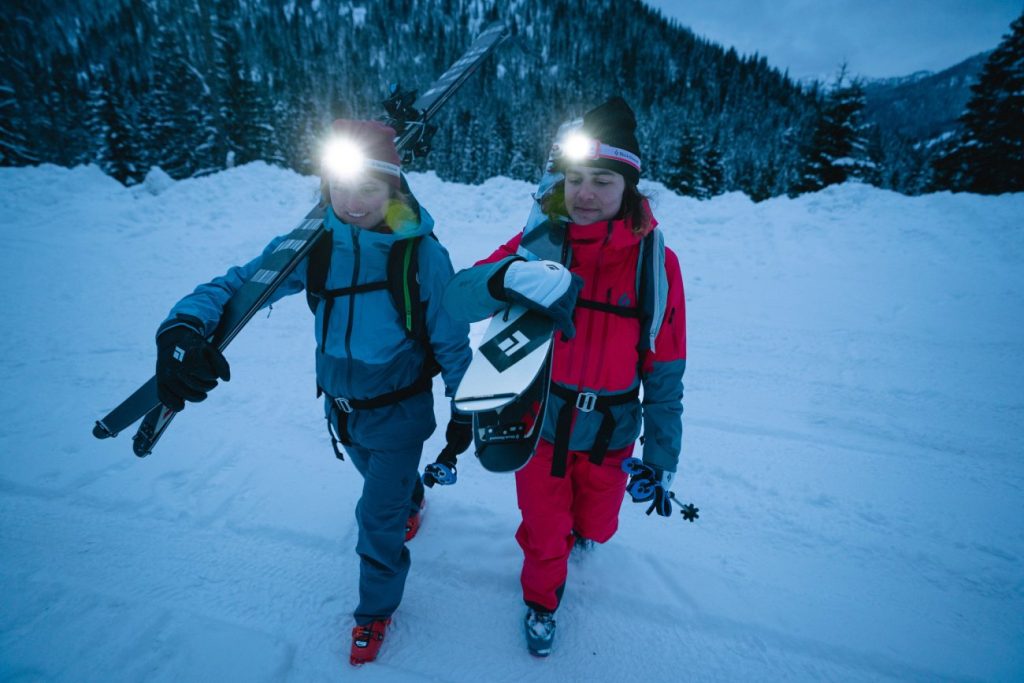 Zwei SkitourengeherInnen mit Stirnlampen, Black Diamond Spark Gloves und geschulterten Ski laufen im Schnee