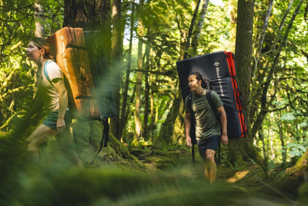 2 Personen mit Crash-Pads auf dem Rücken laufen durch den Wald auf dem Weg zum Bouldern draussen
