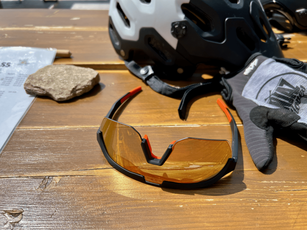 Detailaufnahme der react optray Sonnenbrille auf einem Tisch liegend, Bike-Helm und -Handschuhe im Hintergrund